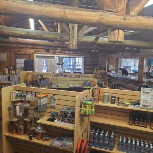 A rustic log cabin gift shop interior featuring shelves with souvenirs, snacks, water bottles, small potted plants, and various items. Wooden beams and natural light create a cozy atmosphere.