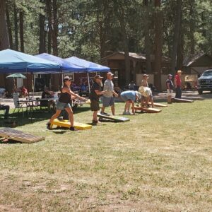 People playing cornhole outdoors under tall pine trees, with several canopies set up for shade and others watching or relaxing in the background at what looks like a campsite.