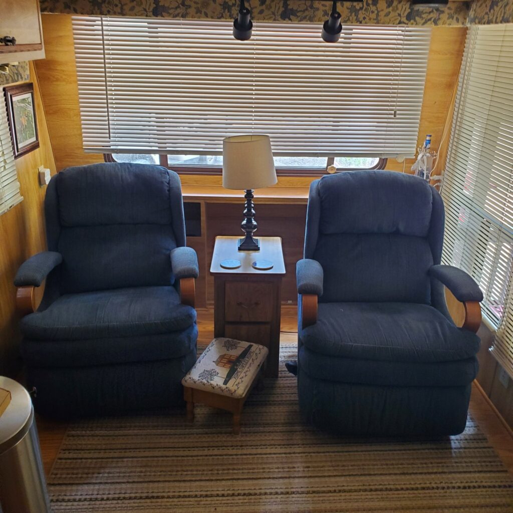 Two blue recliner chairs sit side by side in front of a window with closed blinds in this cozy vacation cabin. A small wooden table with a lamp separates them, and a footstool rests nearby. Wood-paneled walls and a striped rug complete the inviting space.