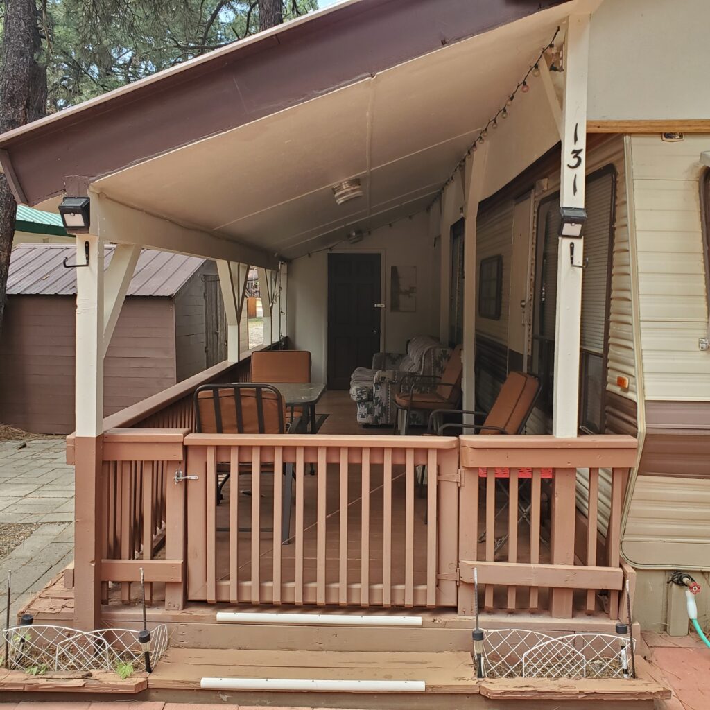 A covered porch with a brown railing, outdoor furniture, and string lights creates a cozy spot for cabin rentals. The porch is attached to a trailer with the number 131 on the post, and two flower boxes sit on the steps leading up.