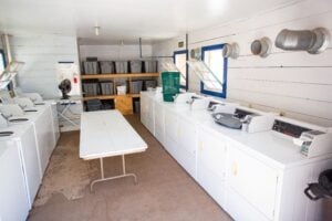 Laundry room with white washers and dryers along the walls, a folding table in the center, storage bins on shelves, and vents on the right wall.