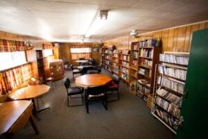 A small library room with wood-paneled walls, bookshelves filled with books and DVDs, tables and chairs arranged for reading or studying, and curtained windows letting in natural light.