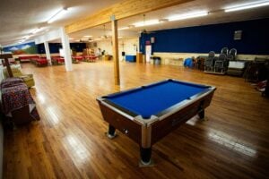 A blue pool table sits on a wooden floor in a large, empty event hall with stacked chairs and round tables in the background.