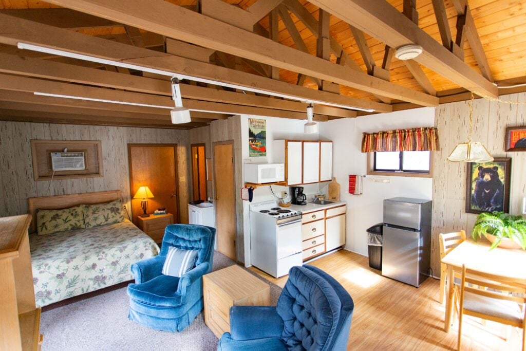 Interior of a cozy cabin with exposed wooden beams, a bed, kitchen area, dining table, two blue armchairs, and natural light coming through a window.