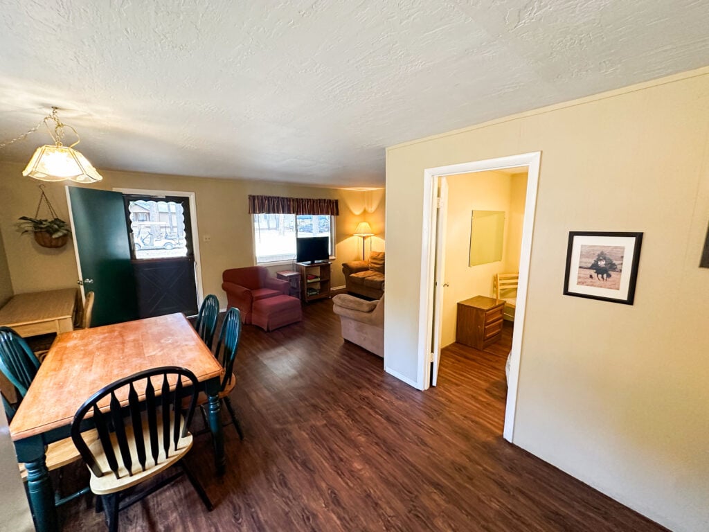 A small living and dining area with a wooden table and chairs, two armchairs, a TV, a lamp, and an open doorway leading to another room. Natural light enters through the window.