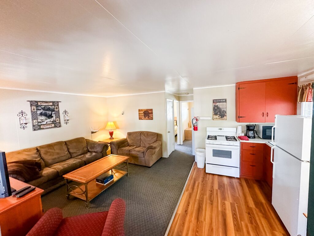 A cozy living room with brown sofas, a coffee table, and a TV, adjacent to a kitchen with white appliances and red cabinets.