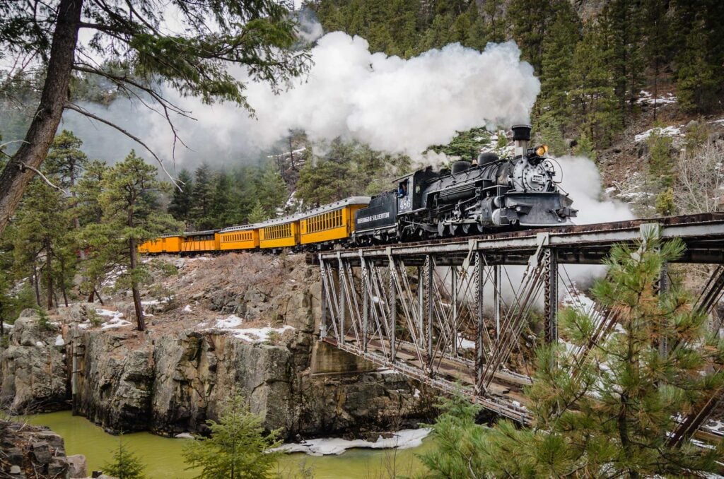 A black steam train with yellow cars crosses a metal bridge over a river, surrounded by pine trees and rocky hills, with white steam billowing from the engine.