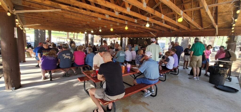 A large group of people gathers under an open wooden pavilion, sitting at picnic tables and eating together on a sunny day.