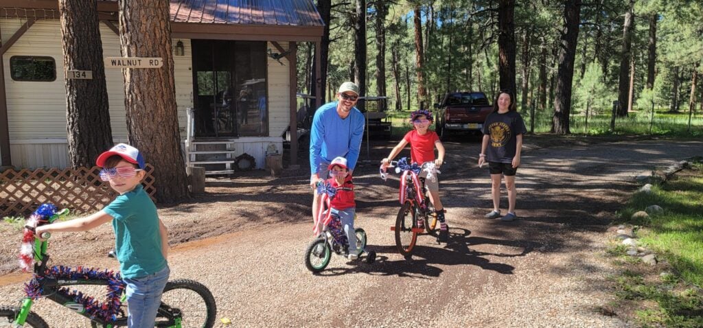 Three children on decorated bikes and one adult stand on a gravel driveway near trees and a house; another child stands nearby, all in casual clothes on a sunny day.