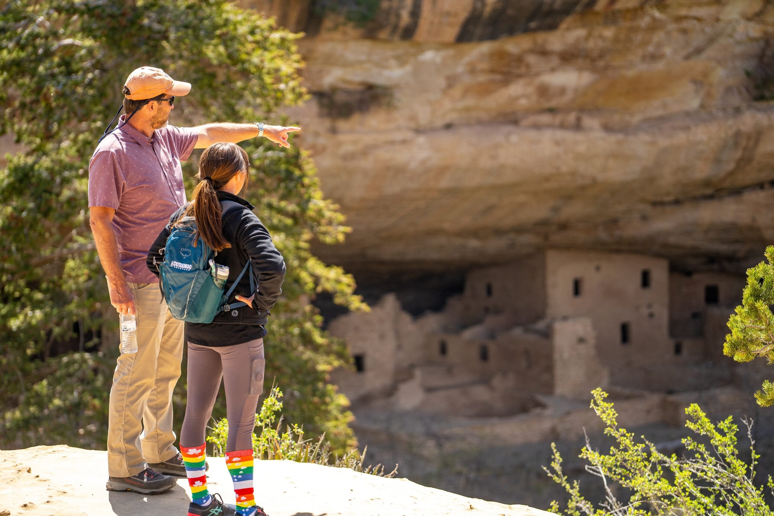 An adult and a child stand on a ledge overlooking ancient cliff dwellings; the adult points toward the ruins while the child observes.