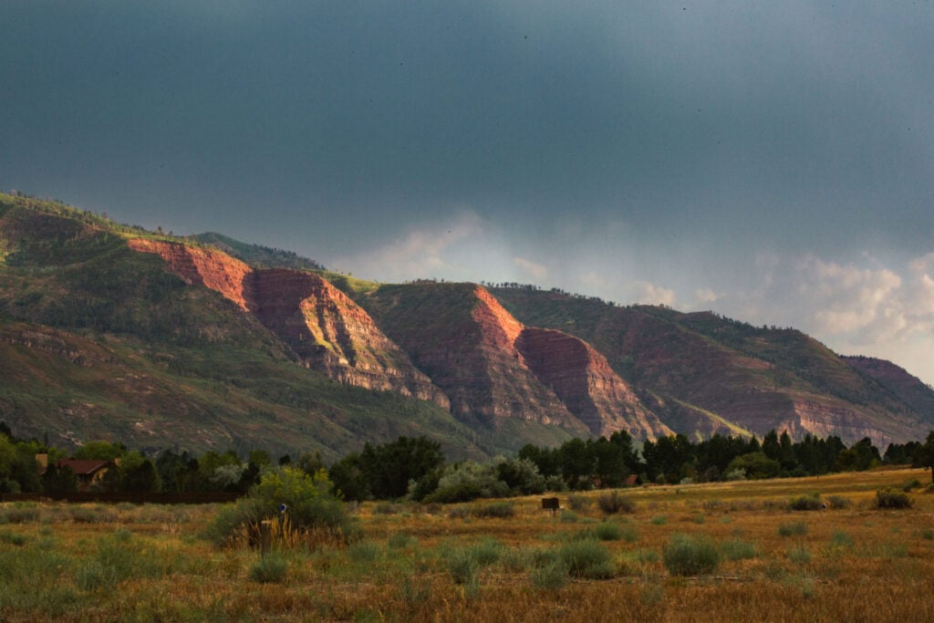 Red rock cliffs are illuminated by sunlight under a cloudy sky, with trees and dry grassland in the foreground.