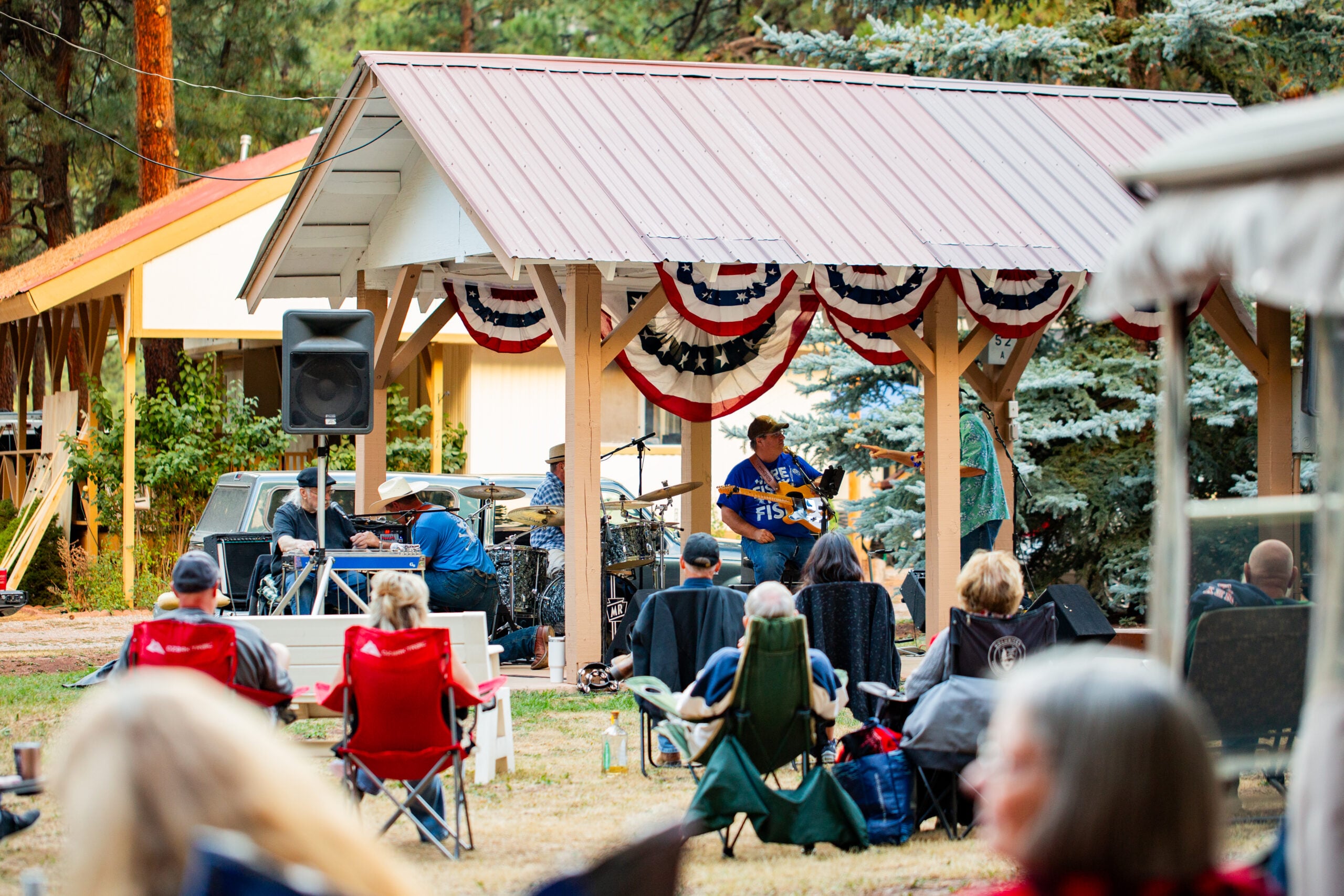 People sit on lawn chairs watching a live band perform under a decorated pavilion at an outdoor event surrounded by trees.