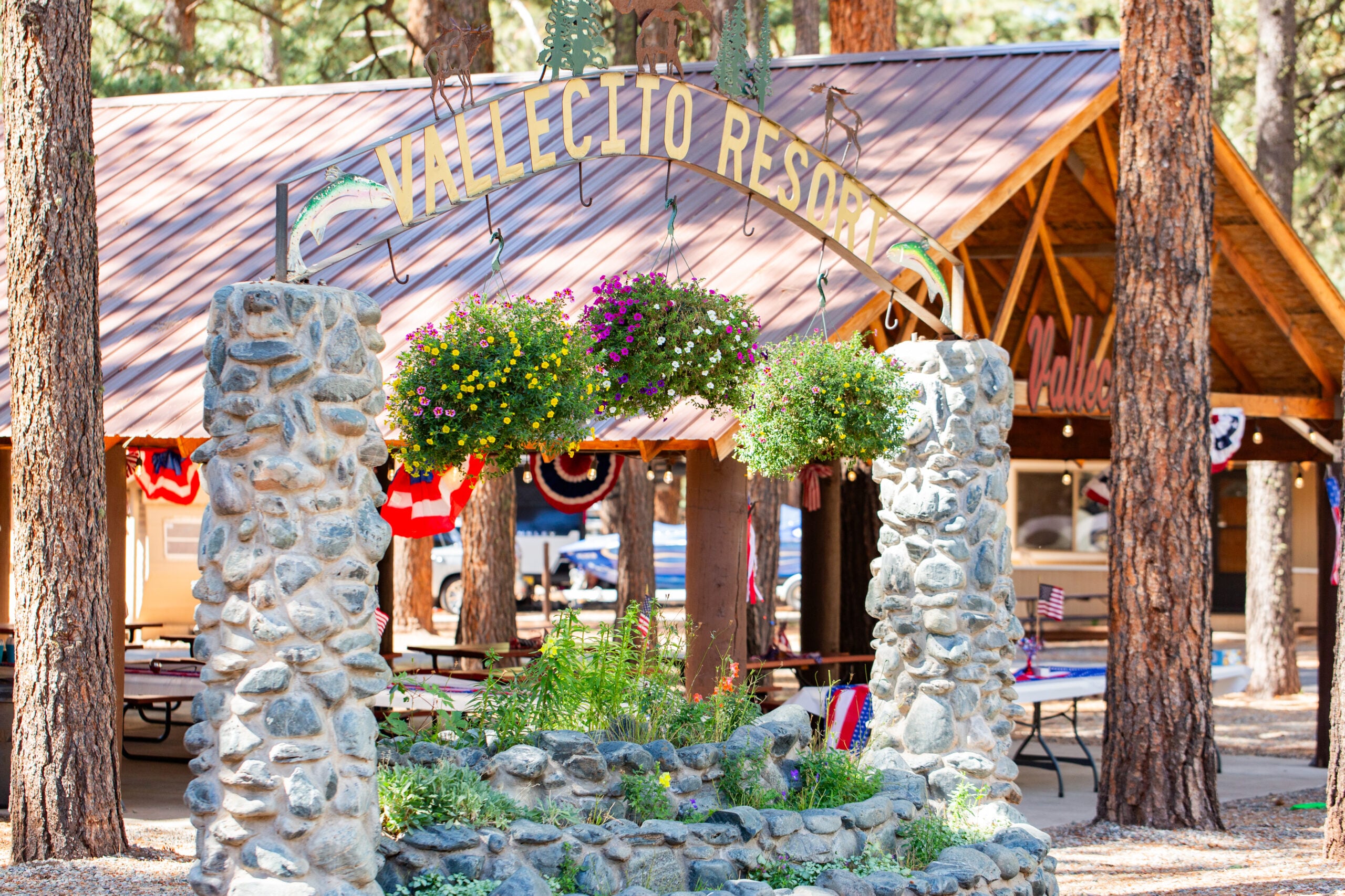 Stone archway with “Vallecito Resort” sign, hanging flower baskets, and picnic area in a wooded setting with wooden pavilion in the background.