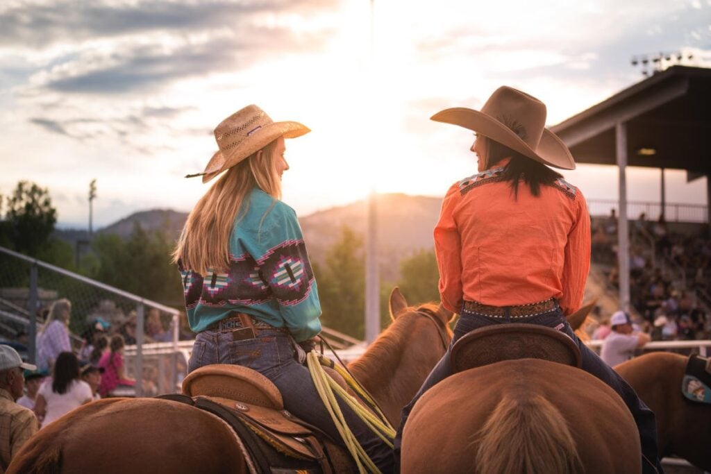 Two people in cowboy hats and western shirts sit on horses at an outdoor rodeo event, facing each other as the sun sets in the background.