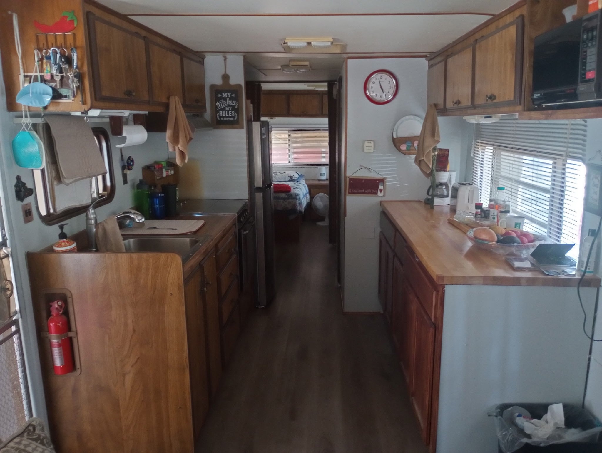 Interior of a camper van with a kitchen, wooden cabinets, countertops, wall clock, and a visible bed area in the back.
