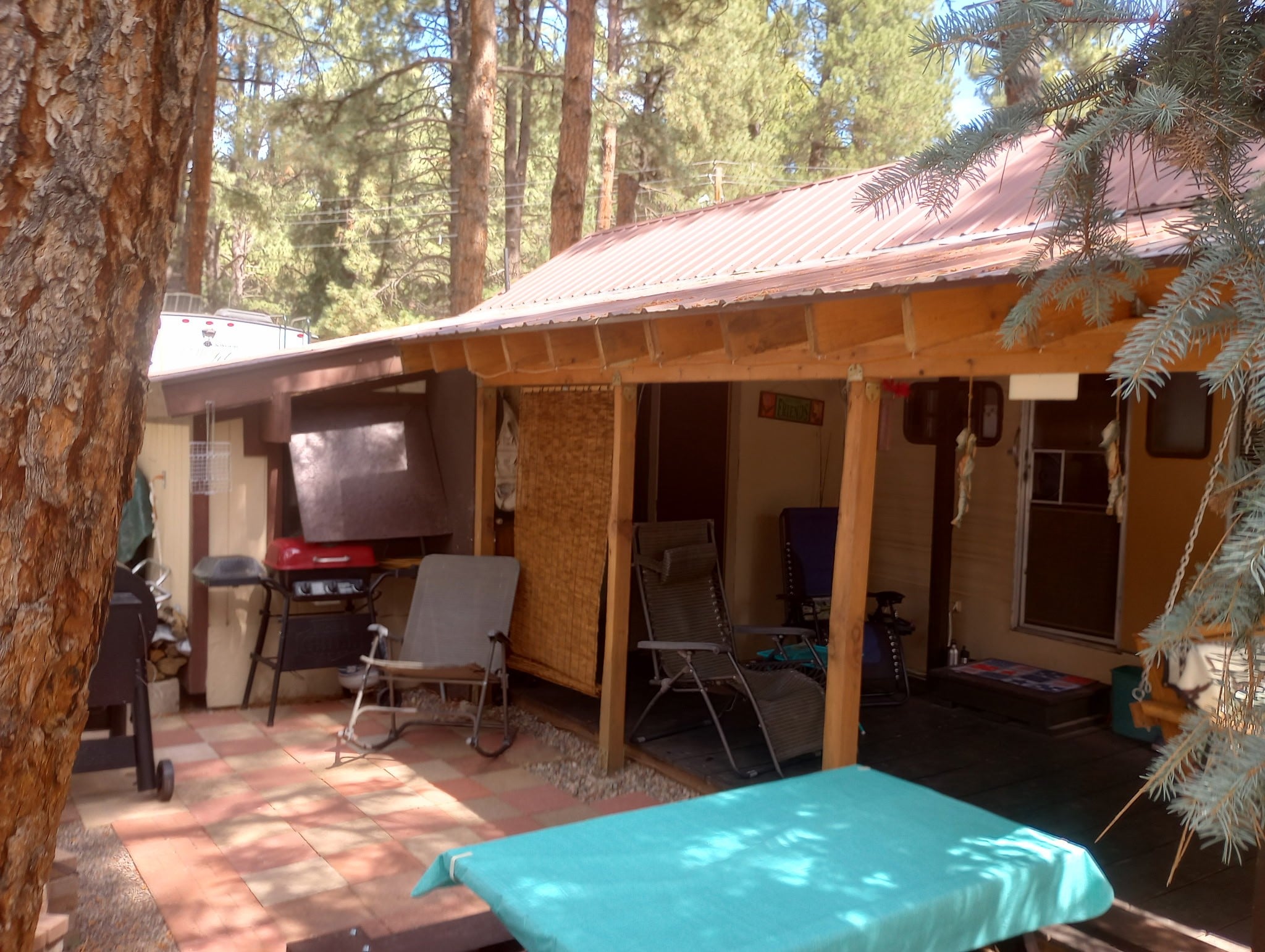 A covered outdoor patio with chairs, a grill, and a table sits next to a tan house surrounded by tall trees in a wooded area.