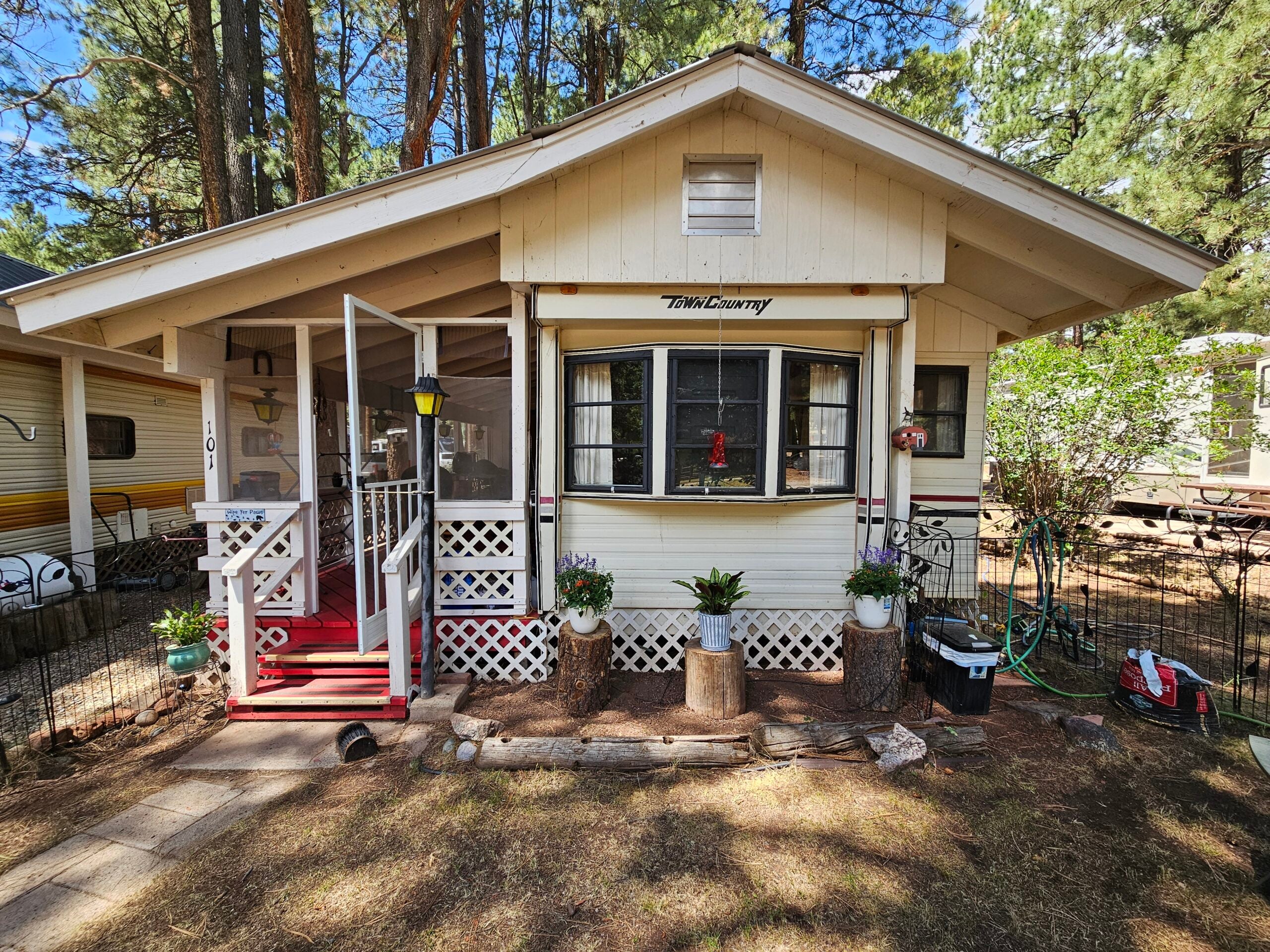 Single-story cabin with a screened porch, potted plants, and lattice accents, surrounded by trees and other trailers in a wooded area.